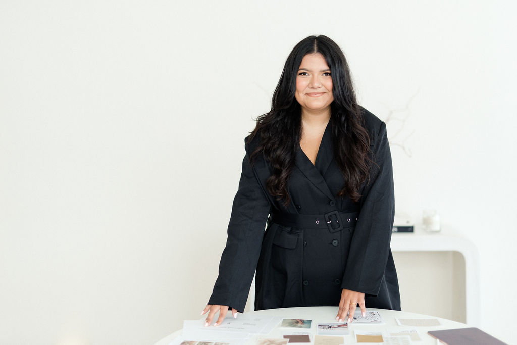 Photo of Christiana Nicole, founder of Briarcroft Lane. Standing at desk in black blazer dress with mood board materials on desk