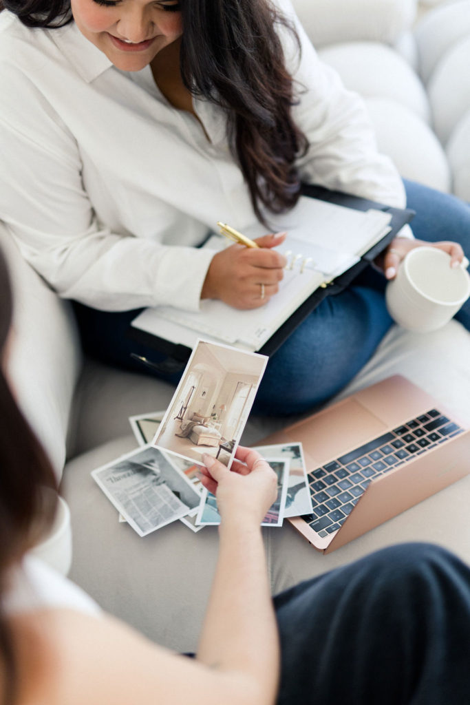 Image of designer sitting with client discussing branding, brand strategy, and website design. Client is holding images while designer is taking notes in a notebook holding a cup of coffee.