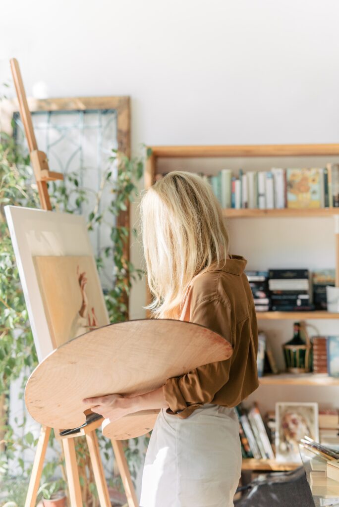 Blonde woman doing a digital detox, holding a painting palette in her left arm while she paints on a canvas with her right hand